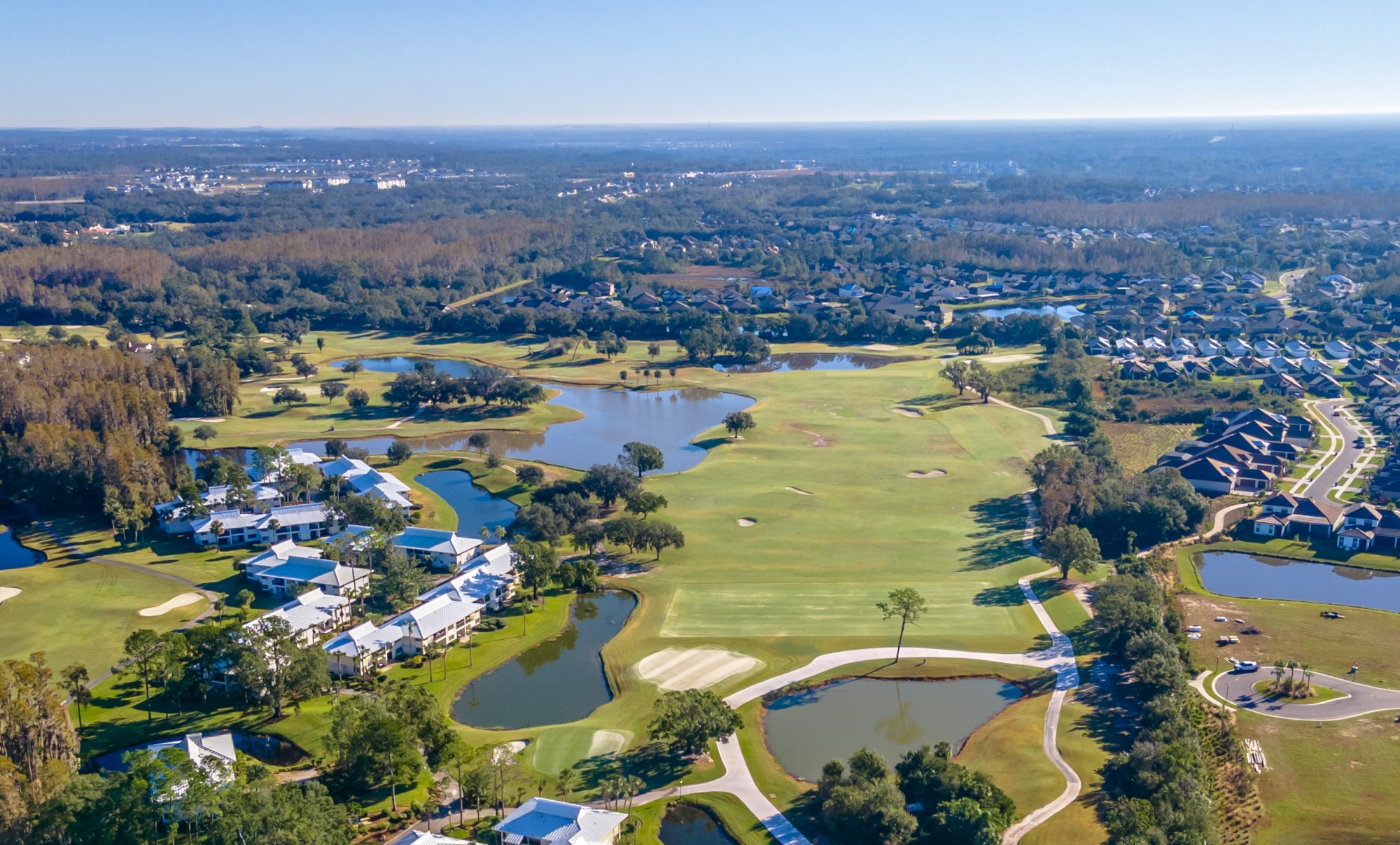 Aerial view of a golf course with several water features, surrounded by lush greenery, houses, and distant landscape under a clear sky.