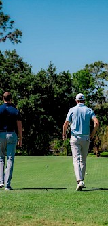 Two people are walking on a green golf course with trees in the background, under a clear blue sky.