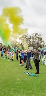 A group of people on a golf course is launching green and yellow smoke, with an audience watching on a cloudy day.