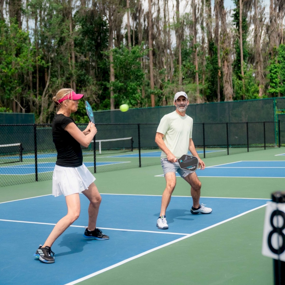 Two people are playing tennis on a court with a green fence in the background.