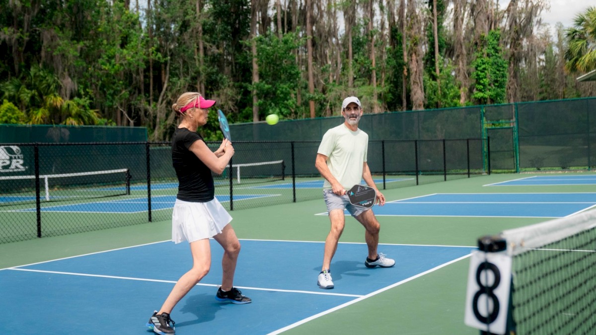 Two people are playing tennis on a court with a green fence in the background.