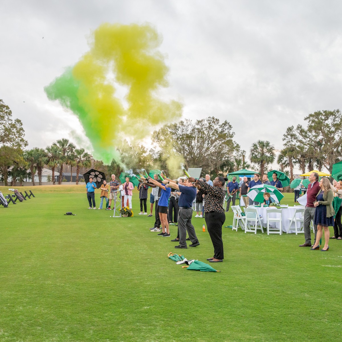 People gathered on a golf course with chairs and green and yellow smoke in the air.