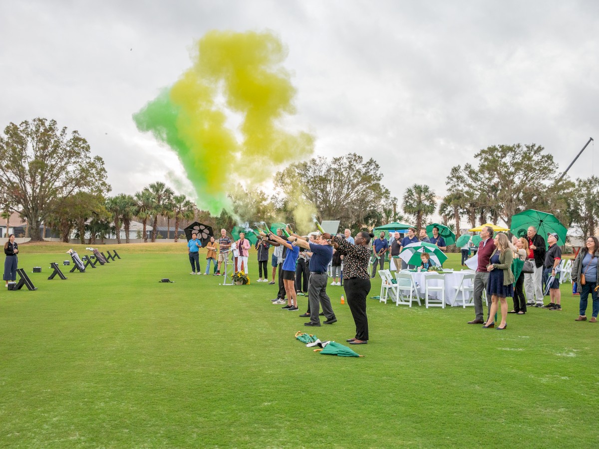 People gathered on a golf course with chairs and green and yellow smoke in the air.