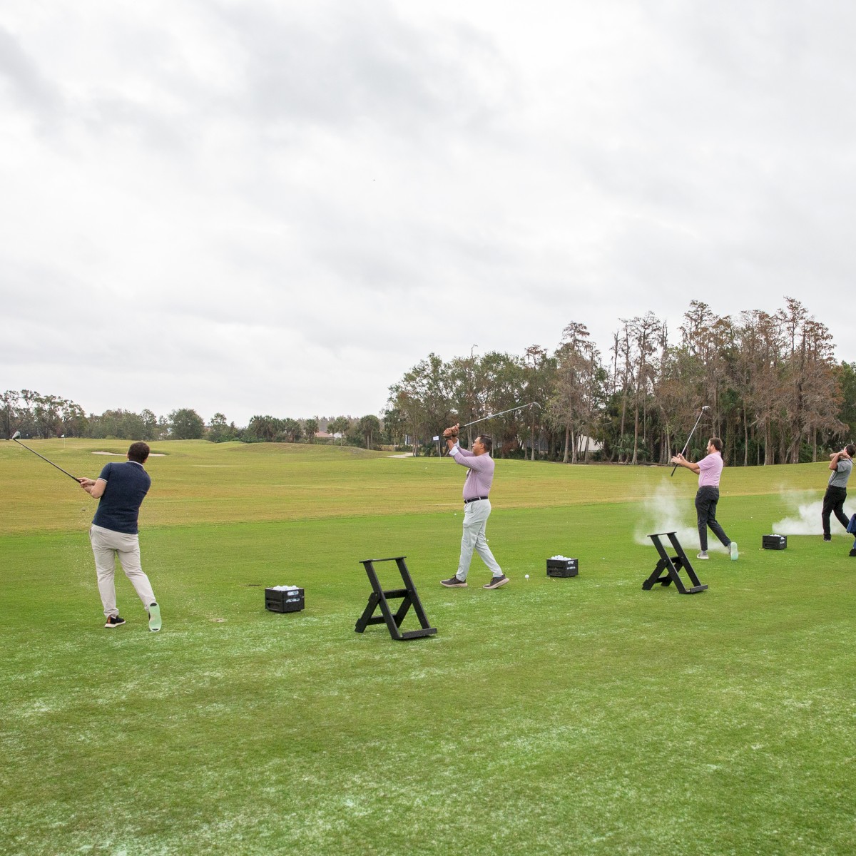 People practicing golf on a driving range with clubs and balls. Green grass, cloudy sky, and trees in the background.