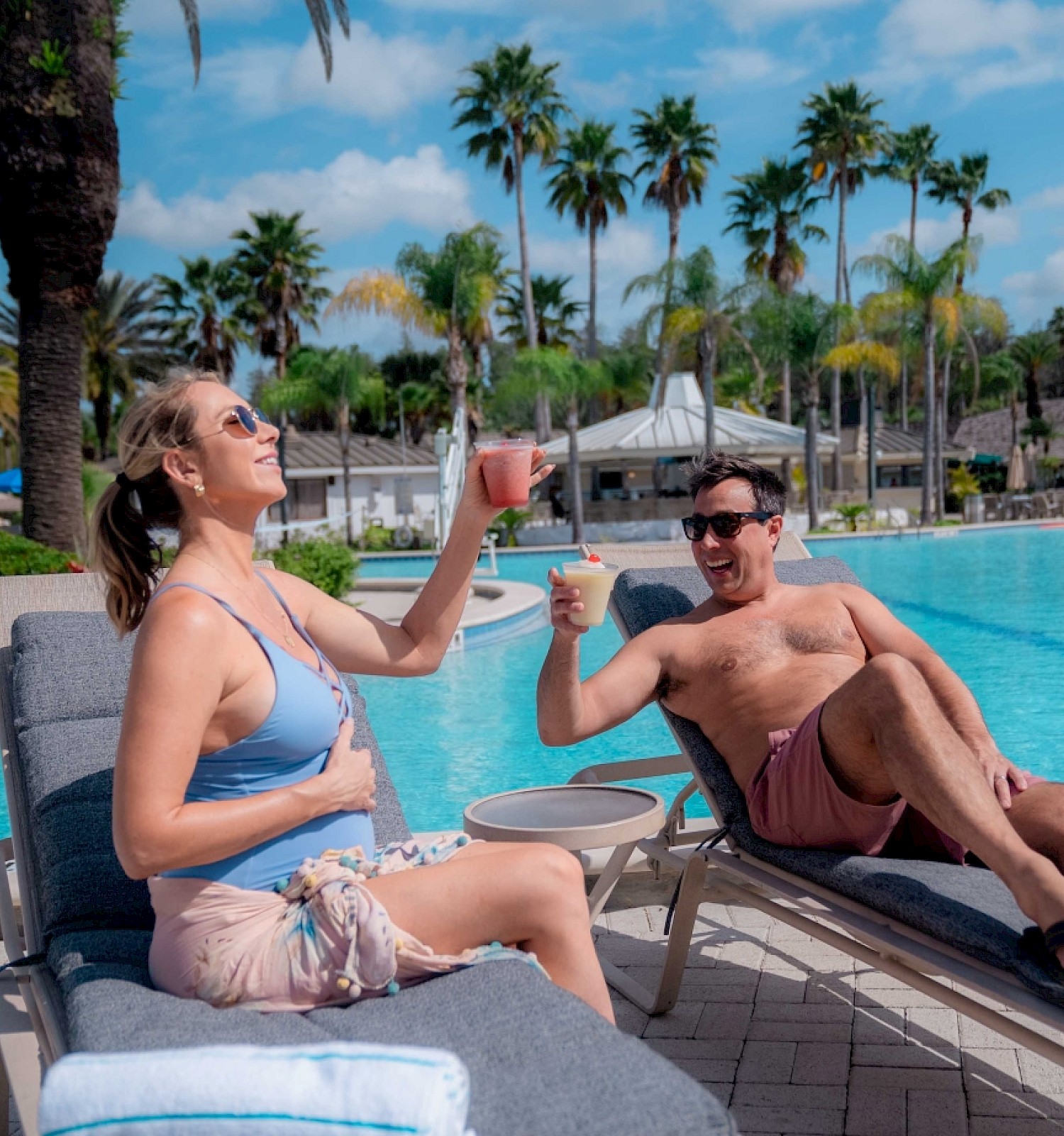 Two people are relaxing by a pool with drinks, enjoying the sunny weather.