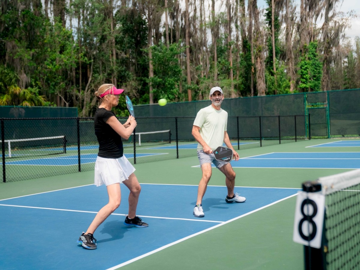 Two people are playing tennis on a court with surrounding greenery.