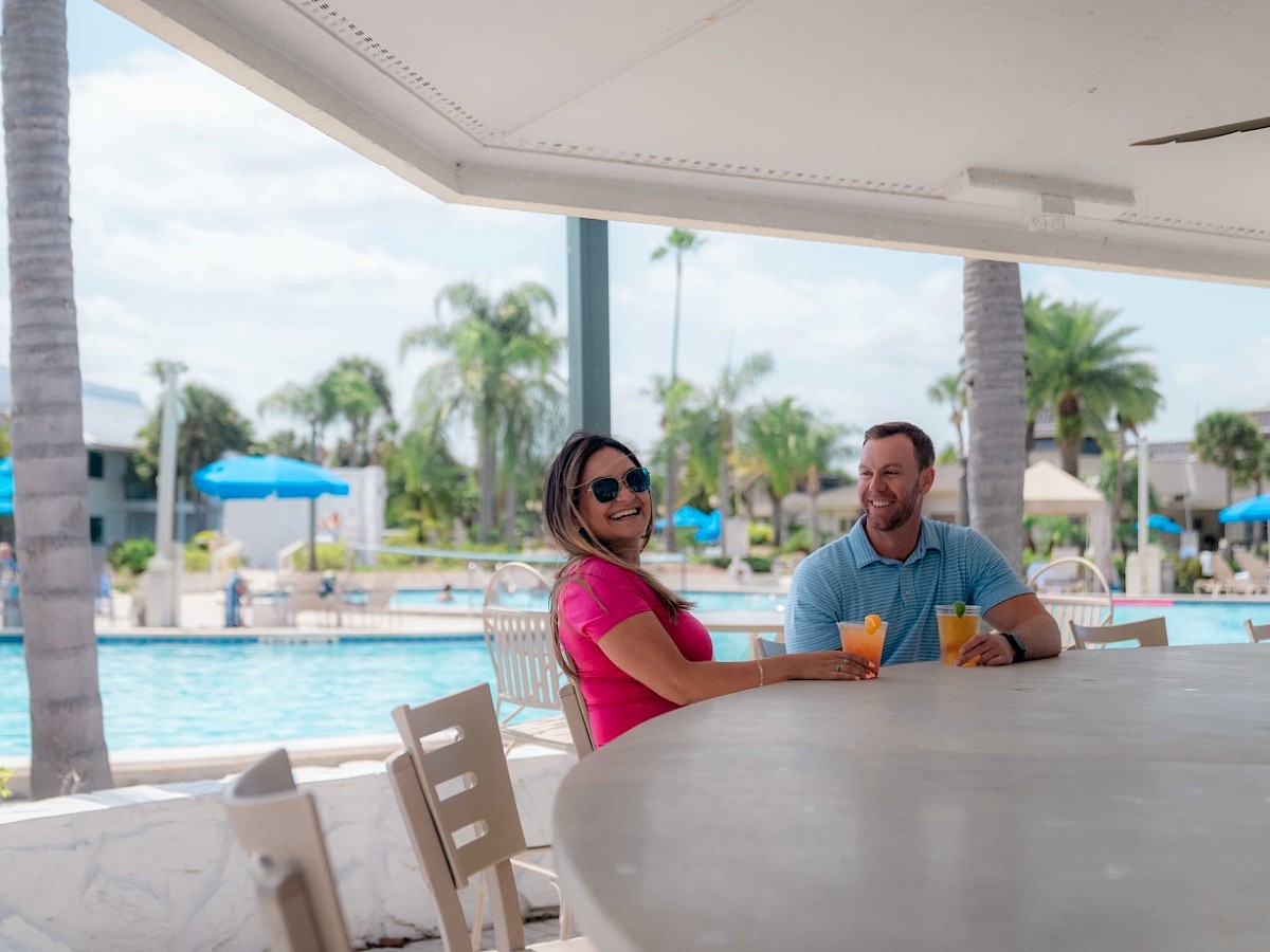 Two people are sitting at a bar by a pool, enjoying drinks and conversation.