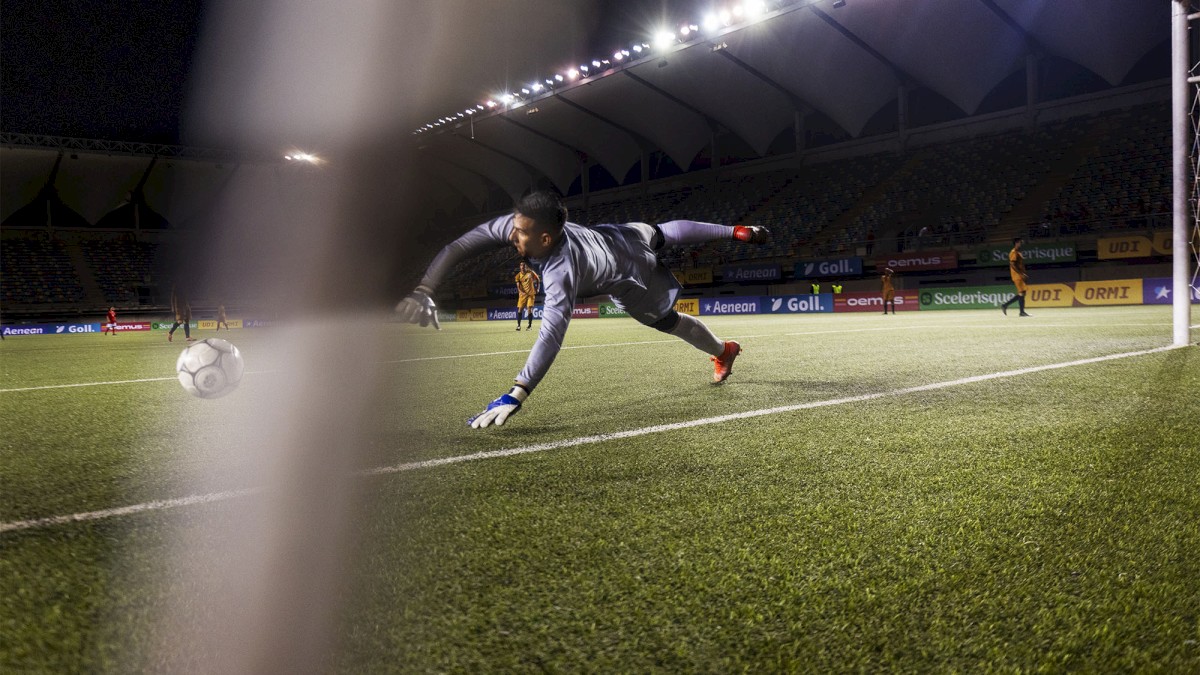 A goalkeeper dives to make a save during a nighttime soccer match, with stadium lights illuminating the field.