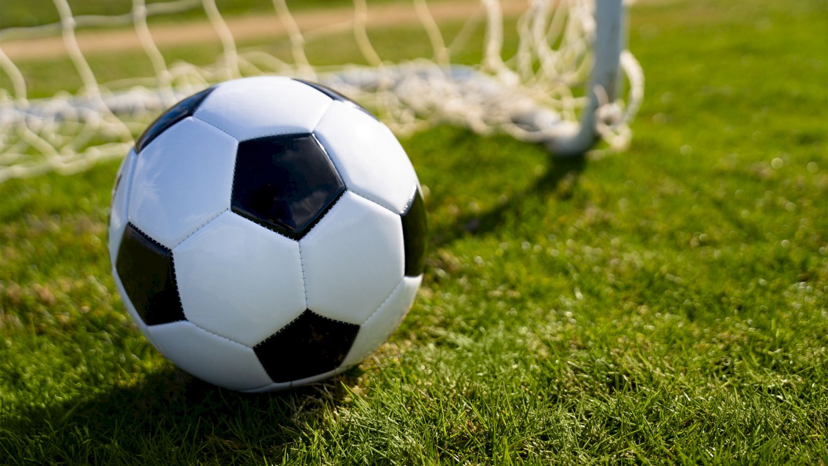 A soccer ball is on green grass near a goal net under clear skies, ready for a game.