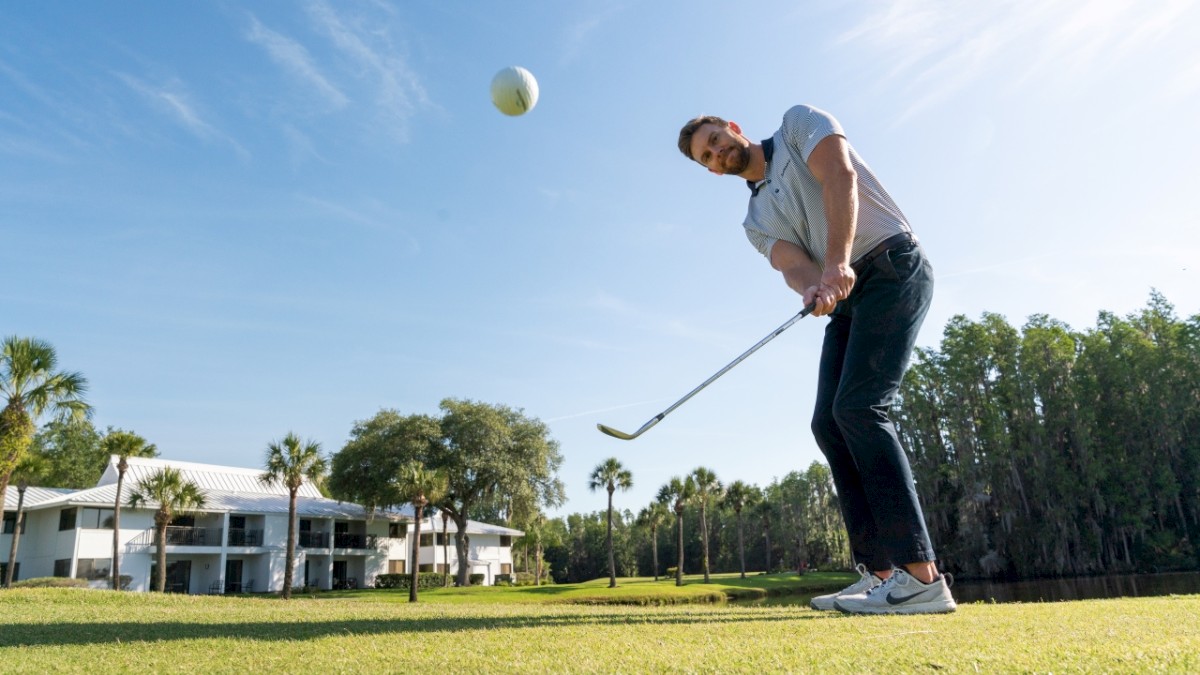 A golfer mid-swing on a lush golf course on a sunny day, with trees and a large building in the background.