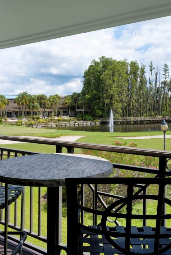 A patio with a table and two chairs overlooks a lush green landscape with trees, a fountain, and a building in the distance.