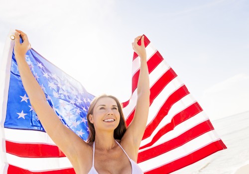 A woman is joyfully holding up an American flag at the beach, with a bright sky in the background.
