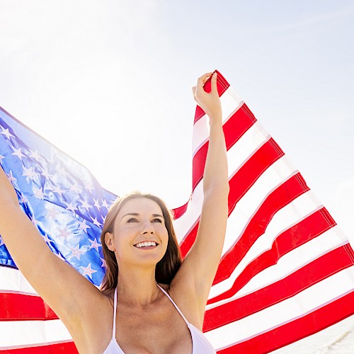 A woman is joyfully holding up an American flag at the beach, with a bright sky in the background.