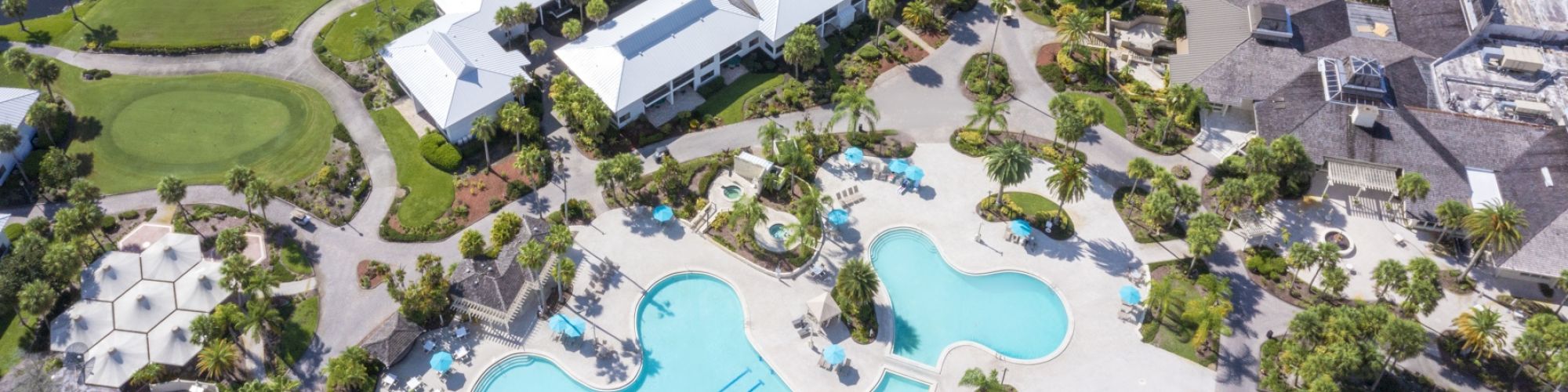 The image shows an aerial view of a resort featuring large swimming pools, white-roofed buildings, landscaped gardens, and water features.