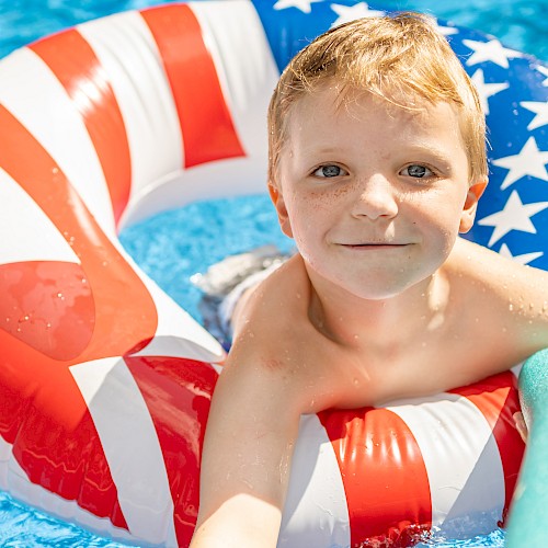 A young boy in a swimming pool is inside a patriotic-themed inflatable ring and holding a green pool noodle, smiling at the camera.
