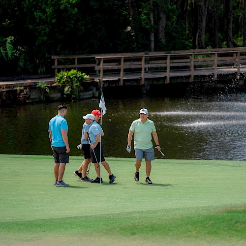 Four people are standing on a golf course near a hole with a flag, next to a pond with a wooden bridge and fountain.