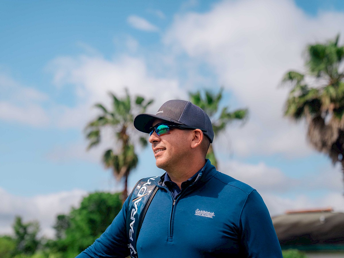 A man in sunglasses and a cap stands outdoors with palm trees in the background, wearing a blue long-sleeve shirt, smiling and looking away.