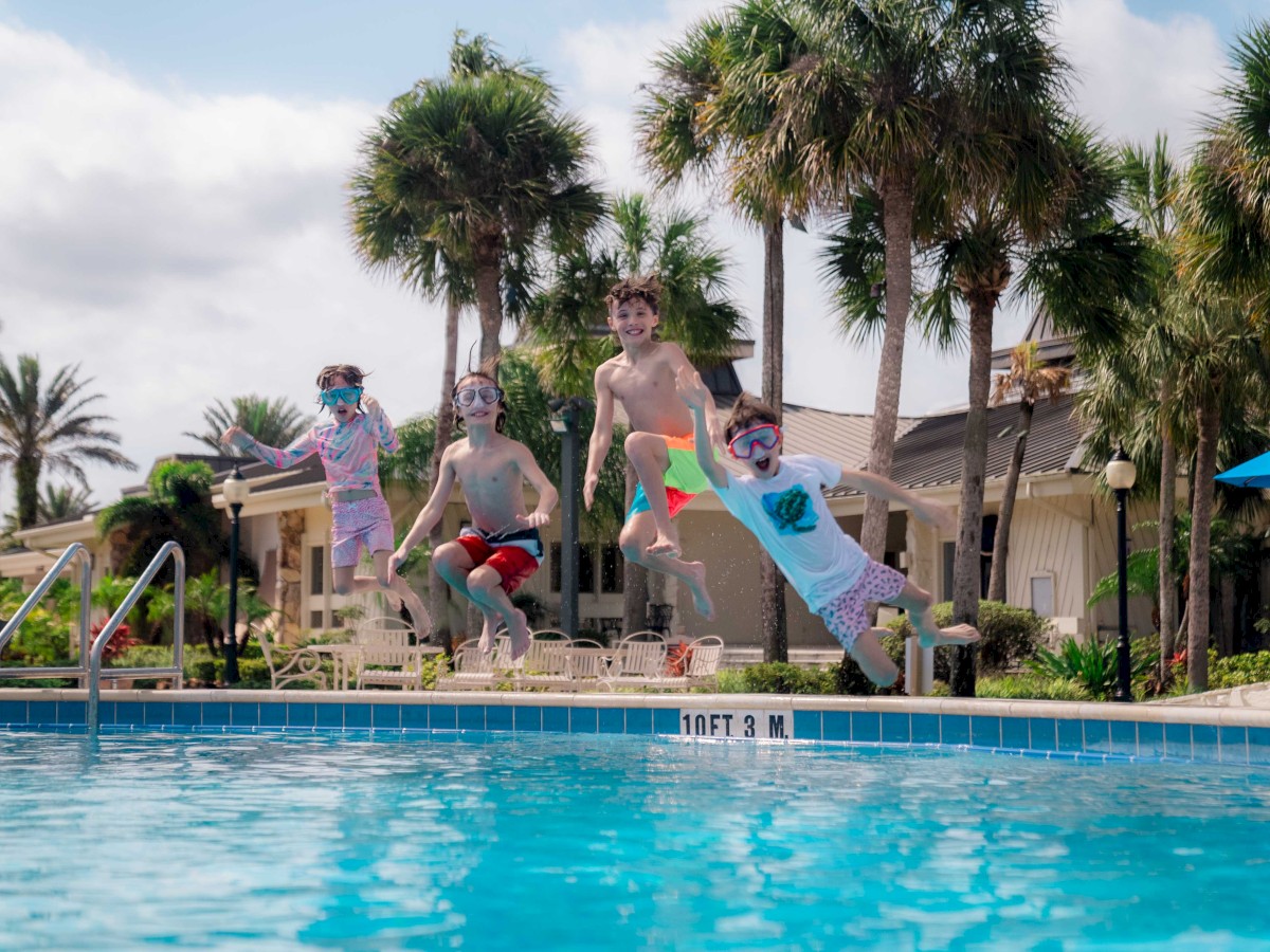 Four children with swim goggles are joyfully jumping into a swimming pool, surrounded by palm trees and resort-like buildings.