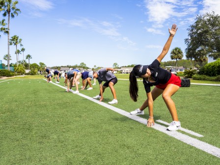 People are stretching on a grassy field, executing side bends in parallel lines under a sunny sky dotted with clouds.