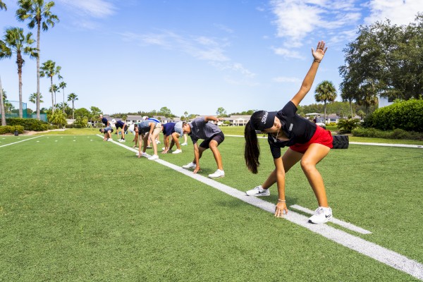 People are stretching on a grassy field, executing side bends in parallel lines under a sunny sky dotted with clouds.