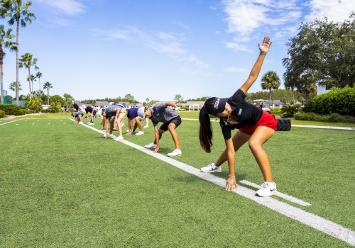 People are outdoors on a grassy field, engaging in a group exercise, stretching while leaning forward and touching the ground.