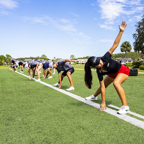 People are outdoors on a grassy field, engaging in a group exercise, stretching while leaning forward and touching the ground.