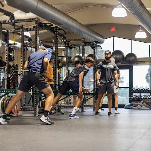 A group of people in a gym engage in exercise and training activities near weightlifting equipment and traffic cones, under a high ceiling.