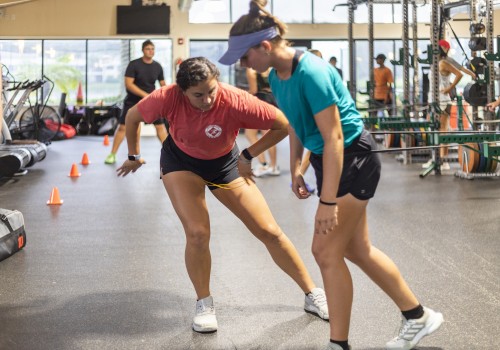 Two people are engaged in a fitness activity in a gym with others in the background, surrounded by gym equipment and training cones.