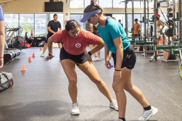 Two women appear to be engaged in a dynamic exercise in a gym, with others working out in the background and cones set up on the floor.