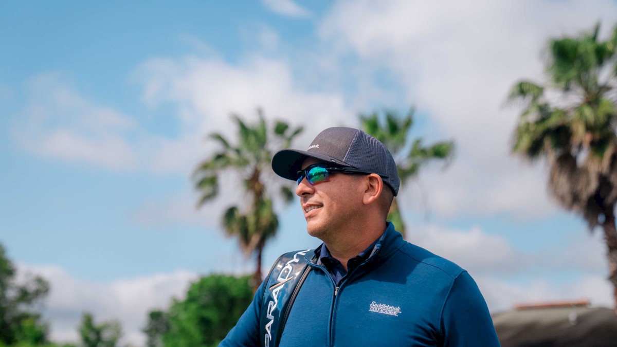 A person wearing a cap, sunglasses, and a blue top stands outdoors with palm trees and a partly cloudy sky in the background, appearing to smile.