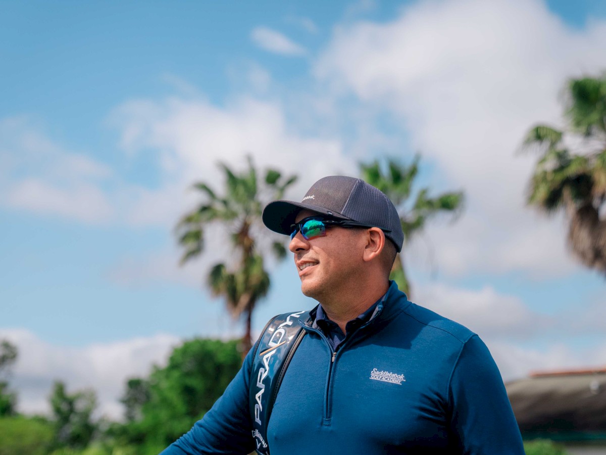 A person wearing a cap, sunglasses, and a blue top stands outdoors with palm trees and a partly cloudy sky in the background, appearing to smile.