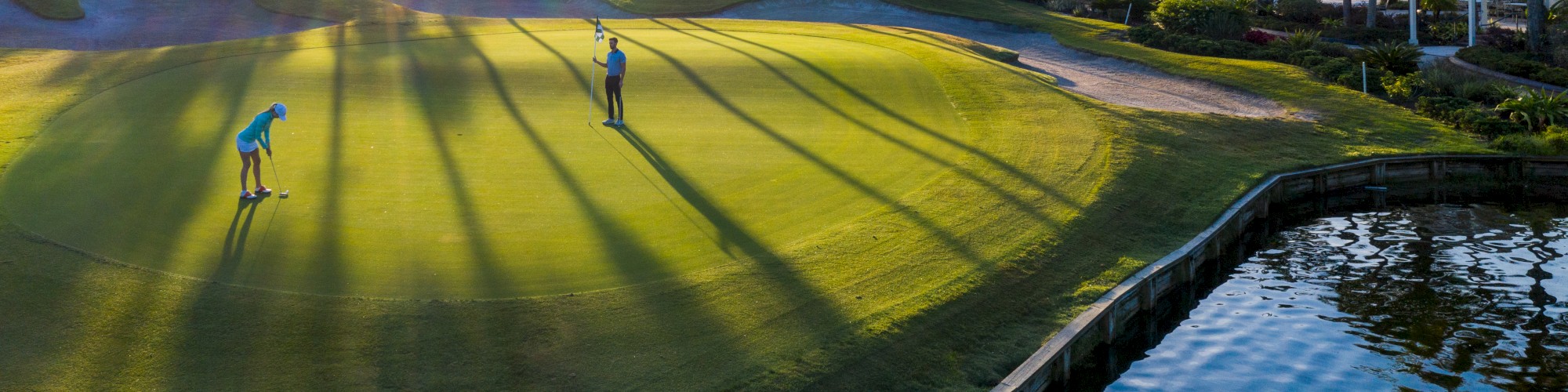 Two people are playing golf on a well-maintained course near a body of water, surrounded by palm trees and a lush setting, at sunset.