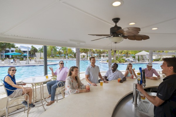 People sitting at an outdoor bar near a pool, being served drinks by a bartender. It’s a sunny and pleasant environment, perfect for relaxing.