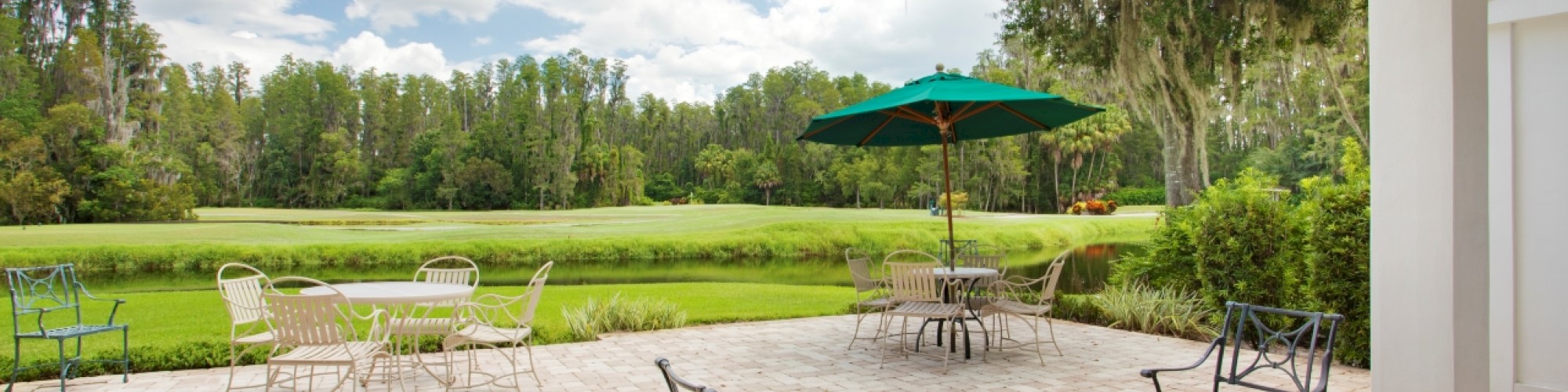 A patio overlooks a lush green landscape with tables, chairs, and a green umbrella under a partly cloudy sky, creating a serene outdoor setting.