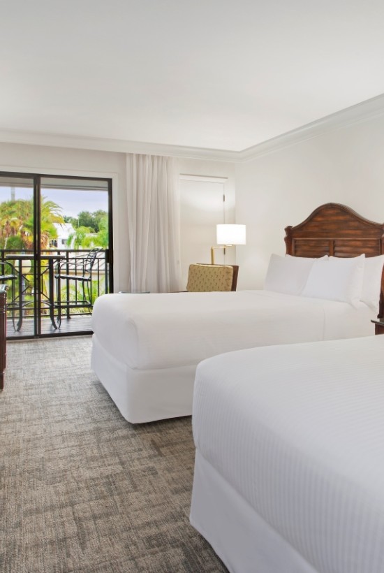 A hotel room featuring two double beds, a TV on a dresser, a desk with a chair, and a balcony with a view of a landscaped outdoor area ending the sentence.