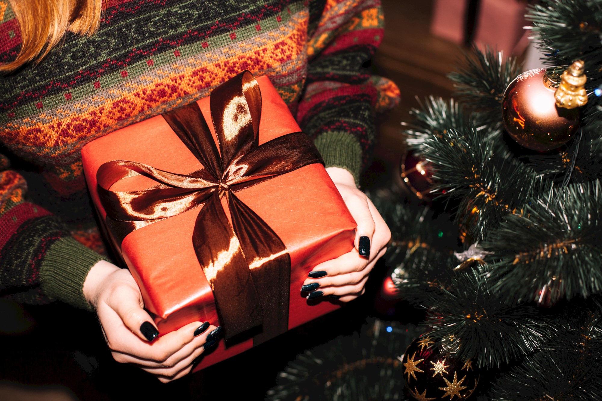 A person in a festive sweater holds a wrapped gift with a bow near a decorated Christmas tree.