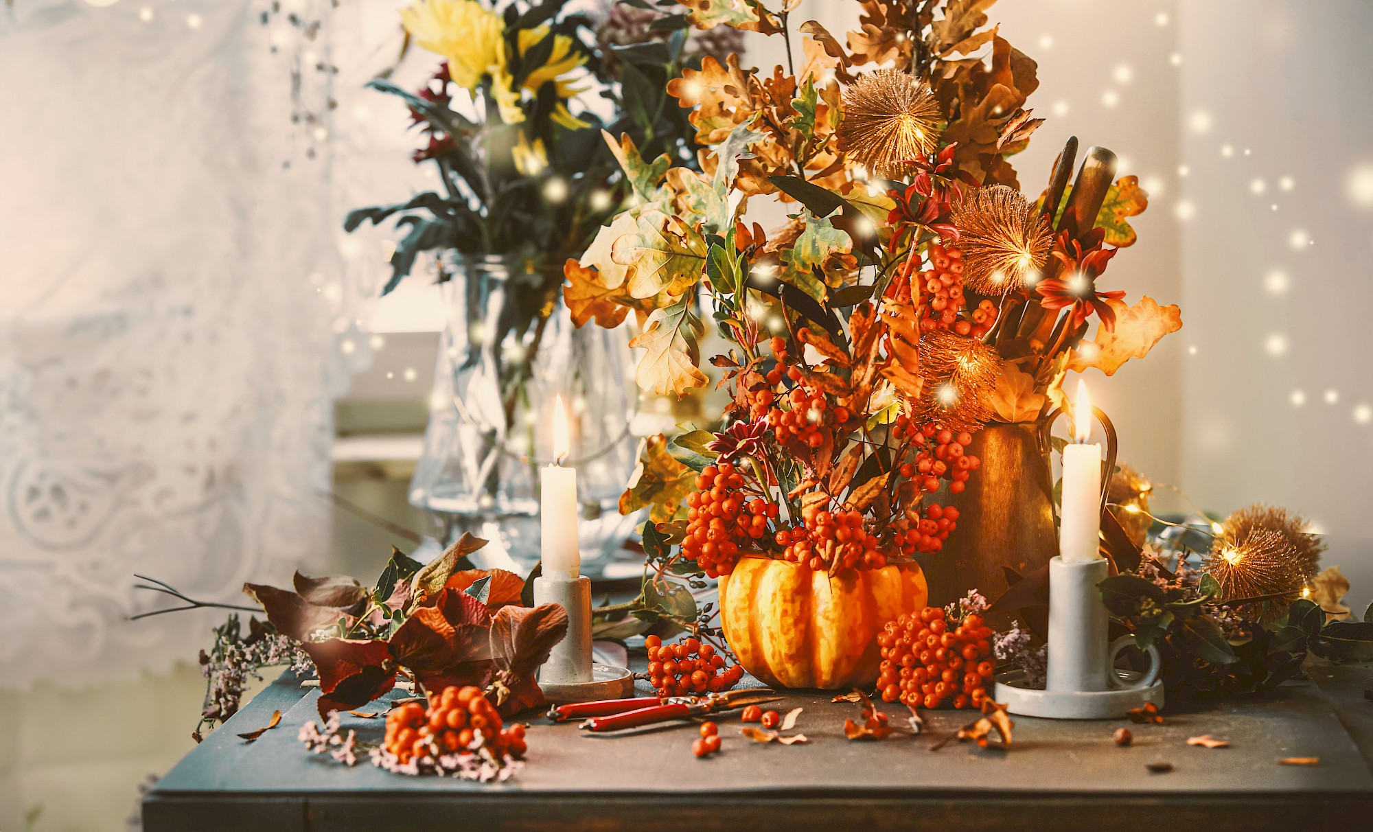 A decorated autumn-themed table includes candles, a small pumpkin, red berries, dried leaves, and flowers, lit warmly with glowing lights.