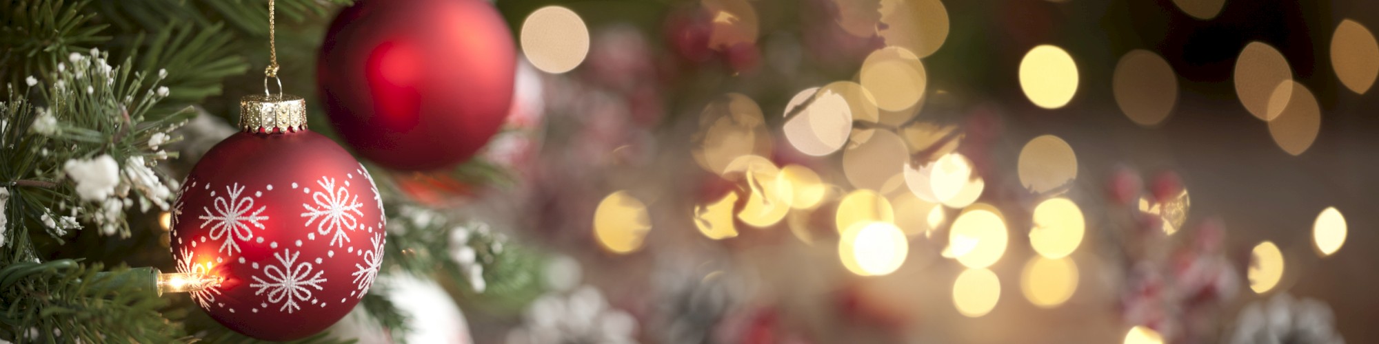 A Christmas tree with red and white ornaments and snowflakes is in focus, while festive lights and decorations blur in the background, creating a bokeh effect.