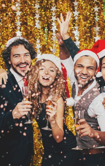 A group of five people wearing Santa hats and holding drinks, celebrating Christmas against a gold, festive backdrop with confetti falling.