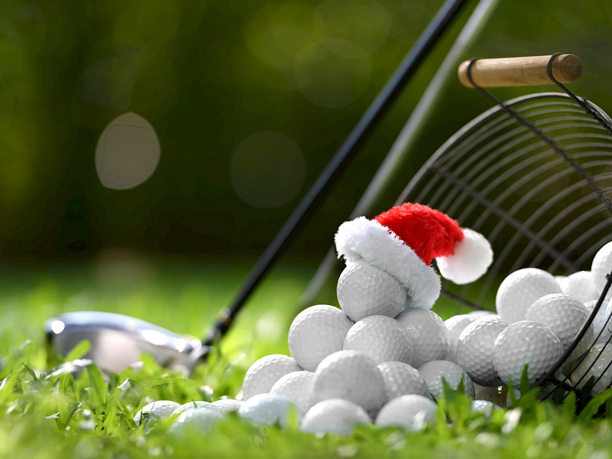 A tipped-over wire basket of golf balls, with one golf ball wearing a small Santa hat, alongside a golf club on a grassy field.