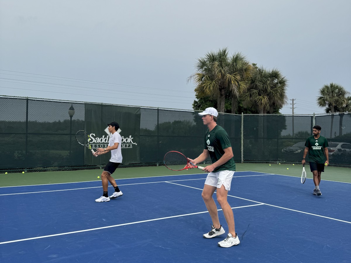 Three people playing tennis on an outdoor court with blue surface and green surroundings, with palm trees and a tennis center sign in the background.