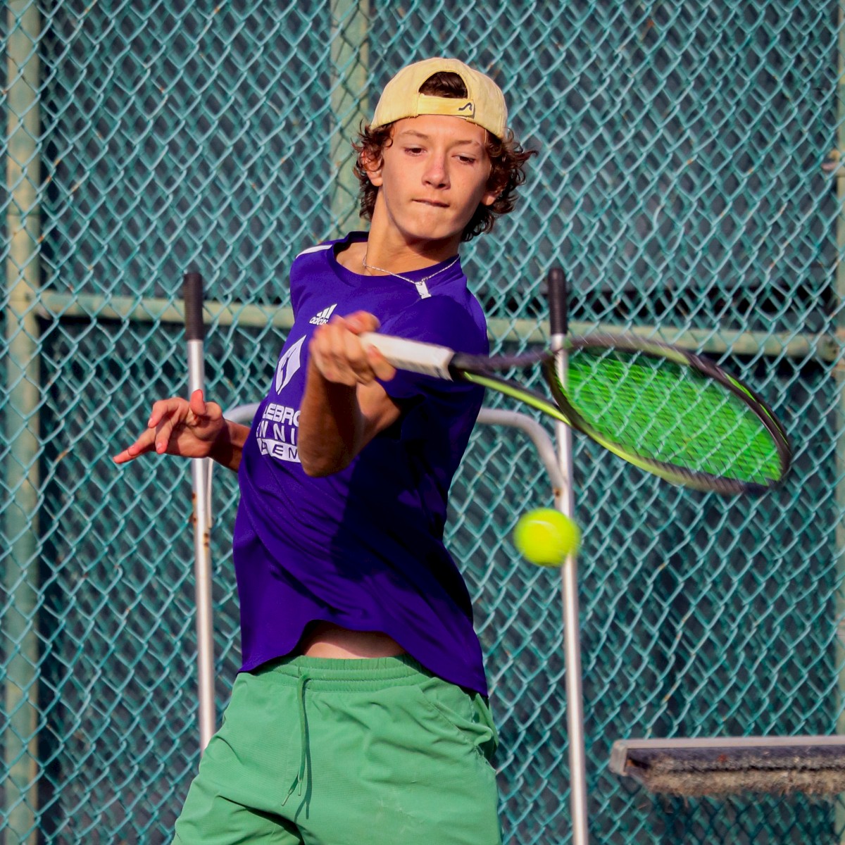 A person in a purple shirt and green shorts playing tennis, focused on hitting the ball with a racket near a chain-link fence.