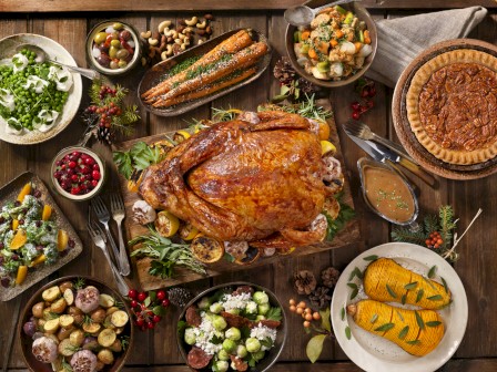 This image shows a Thanksgiving meal with a roasted turkey in the center, surrounded by sides including vegetables, salads, casseroles, and pies.