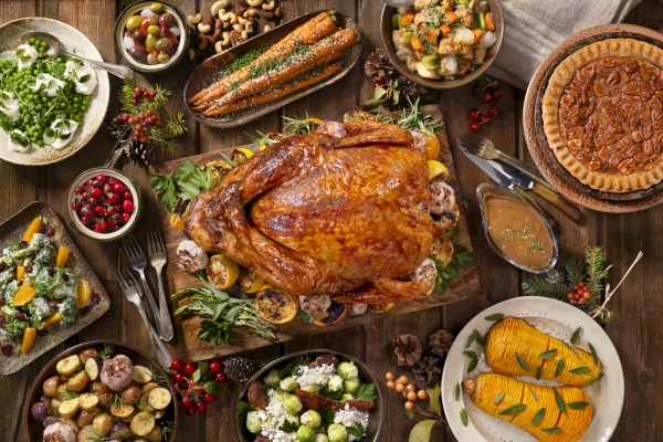 This image shows a Thanksgiving meal with a roasted turkey in the center, surrounded by sides including vegetables, salads, casseroles, and pies.