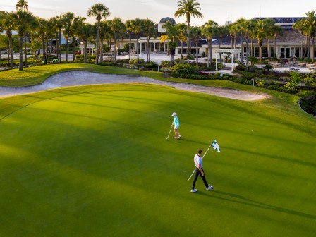 Two golfers on a green course, one holding a flag. Palm trees and a building are seen in the background. The weather appears sunny and clear.