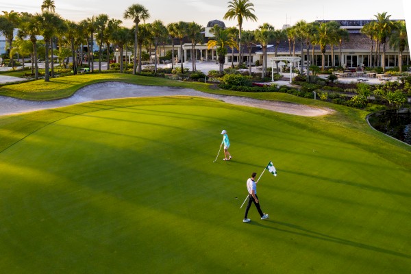 Two golfers on a green course, one holding a flag. Palm trees and a building are seen in the background. The weather appears sunny and clear.