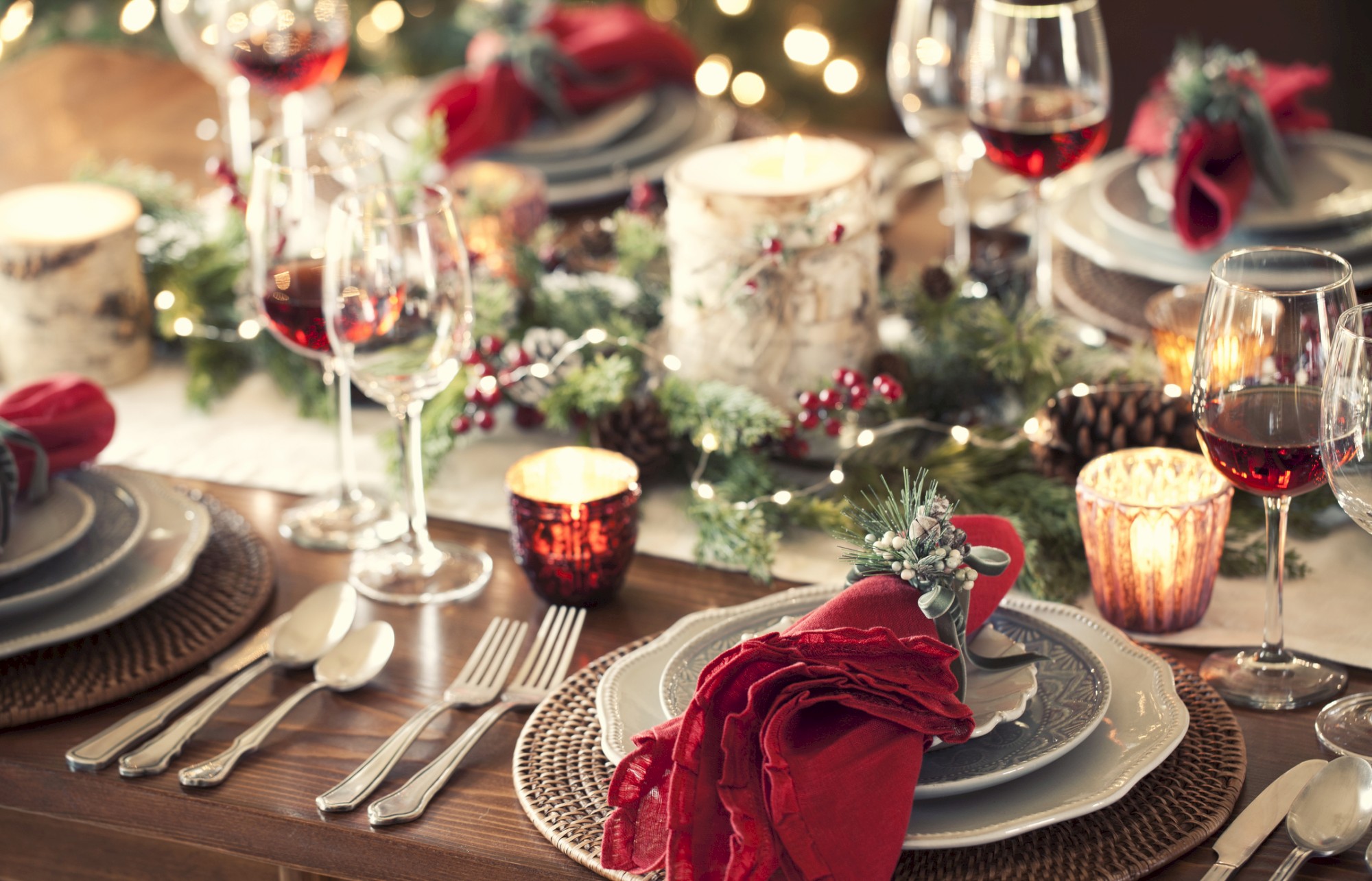 A festive table setting with candles, wine glasses, plates, and silverware, adorned with red napkins and greenery for decoration.