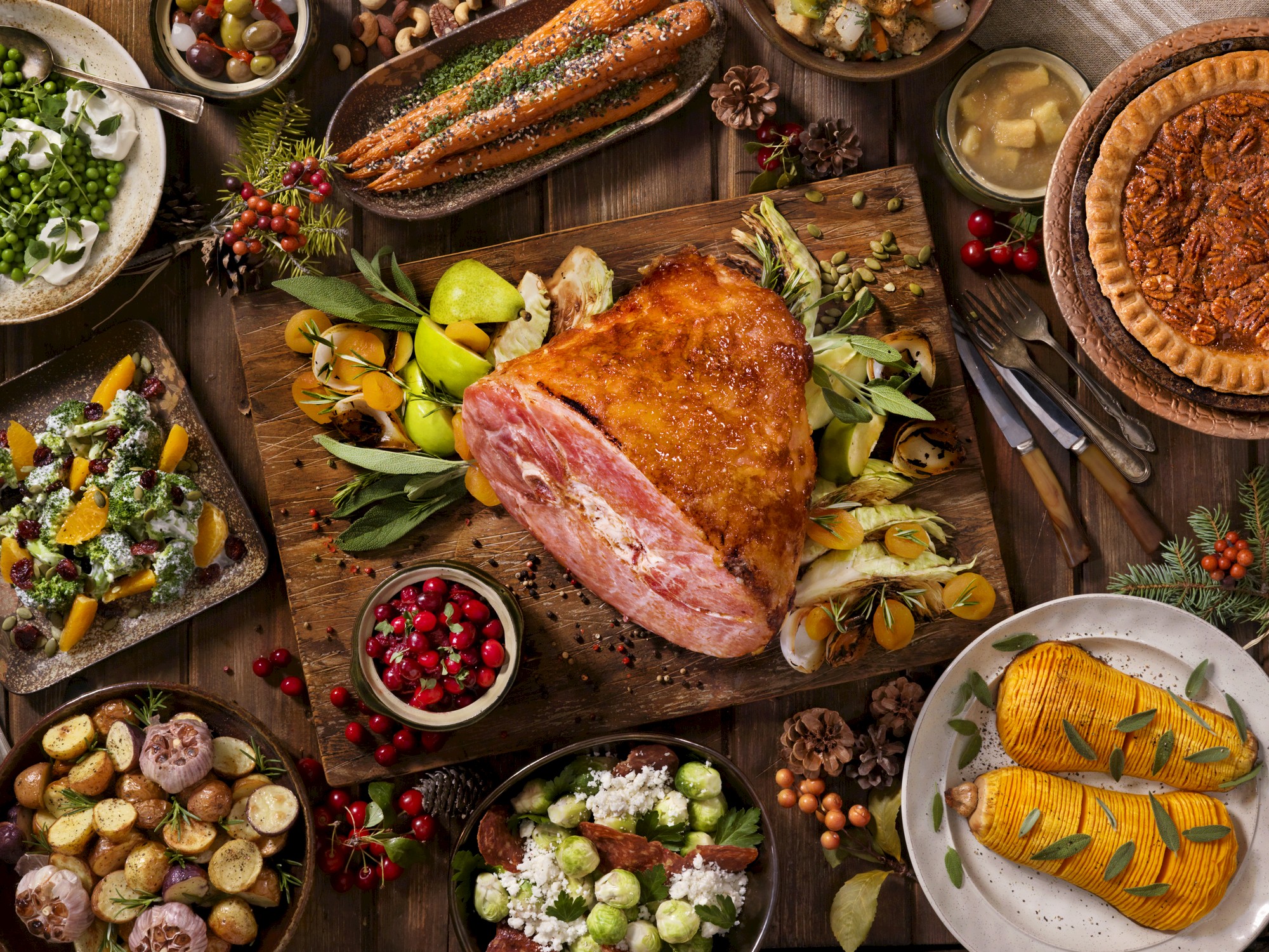 A festive meal spread with ham, vegetables, pie, and various side dishes on a wooden table, perfect for a holiday celebration.