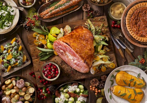 The image shows a festive meal with a central roasted ham, various side dishes, garnished vegetables, and desserts on a wooden table.