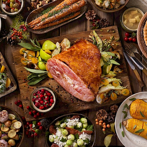 The image shows a festive meal with a central roasted ham, various side dishes, garnished vegetables, and desserts on a wooden table.